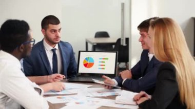 International business partners discuss financial project results on meeting in office. Multiethnic corporate team plans marketing strategy with diagrams, charts, papers, devices. People brainstorming
