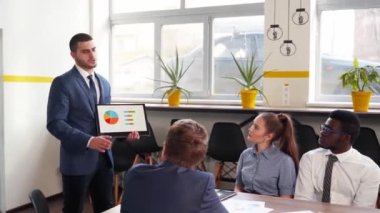 Businessman presents report, shows diagrams on tablet pc in conference room for multiethnic partners. Diverse international team listen CEO on board meeting in office. Corporate people discuss a plan.