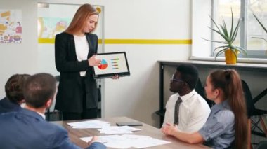 Business woman presents report, shows diagrams on tablet pc in conference room for multiethnic partners. Diverse international team listen CEO on board meeting in office. Corporate people discuss a