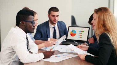 International business partners discuss financial project results on meeting in office. Multiethnic corporate team plans marketing strategy with diagrams, charts, papers, devices. People brainstorming