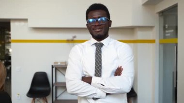 Smiling african american businesman in formal outfit looking at camera standing in an office, arms crossed on chest. Portrait of happy handsome man wearing glasses indoors. Confident corporate worker.