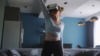 Futuristic girl funny dancing in VR headset for social media platform media in living room at home. Woman avatar performing in metaverse cyberspace immersive experience for her followers audience.