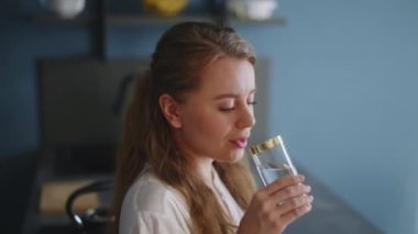 Thirsty smiling young woman standing alone in domestic kitchen drinking water from glass. Girl start a new day with healthy life habit, sipping clean mineral natural still water close up view