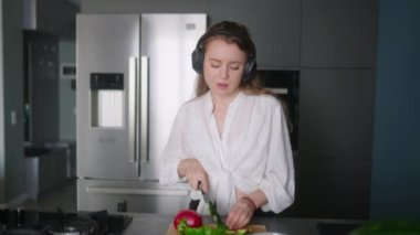 Woman in headphones making a salad of vegetables and has fun dancing at modern kitchen island. Young female cuts the greens and cooks salad. Girl cuts cucumbers and tomatoes on a chopping board.
