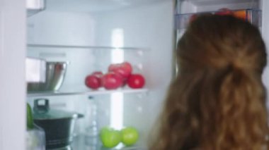 Woman putting groceries in a new modern fridge. Young adult female unpacking shopping bag loading modern hi-tech refrigerator. Girl putting fruit and vegetables into fridge closeup shot