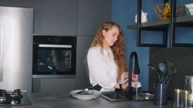 Young caucasian woman washes spoon, fork and knife under running water in sink in modern kitchen. Housewife rinsing cutlery on kitchen island. Female performing the washing-up in high-tech kitchen