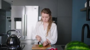 Caucasian woman making a salad of vegetables and tasting a meal on a stove on modern kitchen island. Young female cuts greens and cooks a dish. Girl cuts cucumbers and tomatoes on chopping board.