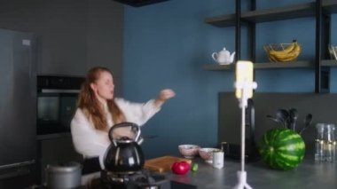 Food blogger woman cooking fresh vegan salad of vegetables in kitchen studio and filming funny dancing for social media content on a smartphone for a video channel. Female influencer streaming.