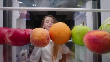 View from inside fridge - woman opens side by side fridge doors, takes fruits and vegetables, closes it. Pretty girl on fruit healthy diet. Female takes tomato, apple, peach, orange from refrigerator.