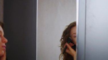 Authentic caucasian woman puts on makeup in front of mirror sitting on the floor. Girl getting ready for work. Young blonde female applying bronzer using big soft fluffy angled brush closeup shot