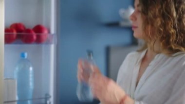 Caucasian woman opens a refrigerator and takes bottle of water, drinks cooled water. Young adult female taking sip of cold water. Girl keeps herself hydrated standing in modern kitchen closeup
