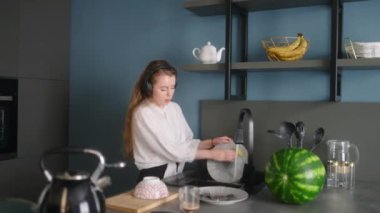 Authentic happy caucasian woman in a headphones washing the dishes and dancing in a modern kitchen. Young housewife listening music, having fun while doing the washing-up.