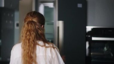 Young woman taking vegetables from fridge and has fun singing and dancing in modern minimalistic kitchen with island. Girl getting tomato from side by side refrigerator and singing to a cucumber.