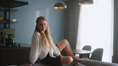 Attractive woman looking at camera sitting on couch on the dark modern stylish kitchen background. Close-up face portrait of young smiling woman. Blonde long-haired female sitting on back of sofa