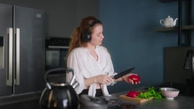 Woman in headphones making a salad of vegetables and has fun dancing at modern kitchen island. Young female cuts the greens and cooks salad. Girl cuts cucumbers and tomatoes on a chopping board.