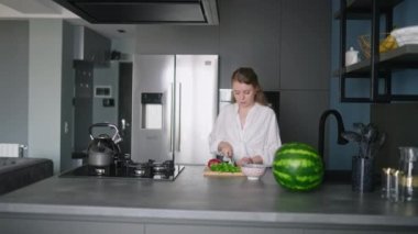 Caucasian woman making a salad of vegetables on modern kitchen island. Young female cuts the greens and cooks a salad. Girl cuts cucumbers and tomatoes on chopping board in minimalistic interior.