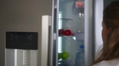 Woman putting groceries in a new modern fridge. Young adult female unpacking shopping bag loading modern hi-tech refrigerator. Girl putting fruit and vegetables into fridge closeup shot
