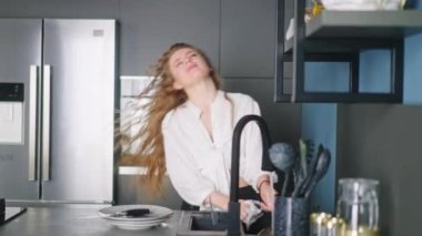 Authentic happy caucasian woman washing dishes and dancing in a modern kitchen. Young housewife having fun while doing the washing-up. Girl waving her hair while doing the dishes.