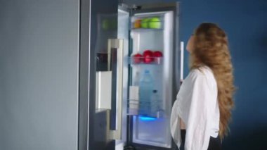 Caucasian woman opens a refrigerator and takes bottle of water, drinks cooled water. Young adult female taking sip of cold water. Girl keeps herself hydrated standing in modern stylish kitchen