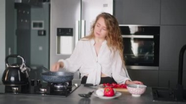 Young curly woman puts tomatoes on hot pan to fry them with scrambled eggs. Young female cooking omelette on gas stove. Girl preparing her breakfast at modern minimalistic kitchen island.