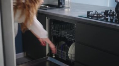 Caucasian woman is unloading clean plates from a dishwasher machine in modern stylish high-tech kitchen. Young pretty housewife opens dishwasher, takes out dishes then closes it