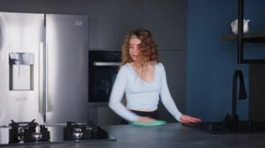 Pretty young caucasian woman wearing headband wipes surfaces at home in a modern stylish kitchen, keeping house neat. Housewife cleaning kitchen surface with rag. Young female doing household chores