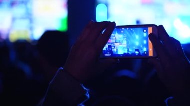 Close up of person hands silhouette taking photo or recording video of live music concert with smartphone at open air festival. Bright colorful stage lighting. Fans at the favourite band performance.