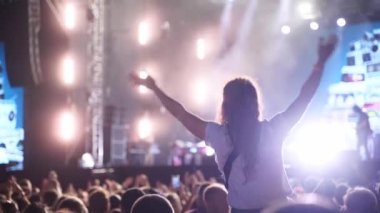 Woman sits on boyfriend shoulders clapping hands at music concert on open air fest. Crowd applause, jump, wave hands and dance at a favourite band performance on stage. Bright illumination.