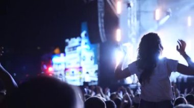 Woman sits on boyfriend shoulders waving hands at music concert on open air fest. Crowd applause, jump, wave hands and dance at a favourite band performance on stage. Bright illumination.
