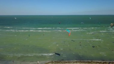 Aerial of many kiteboarders with colorful kites flying over the blue sea lagoon ride on kiteboards. Kitesurfers surf on waves with parachutes on a windy day. Drone video.