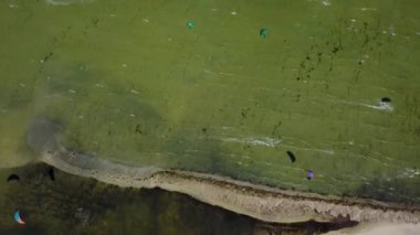 Aerial of many kiteboarders with colorful kites flying over the blue sea lagoon ride on kiteboards. Kitesurfers surf on waves with parachutes on a windy day. Top view from above video.
