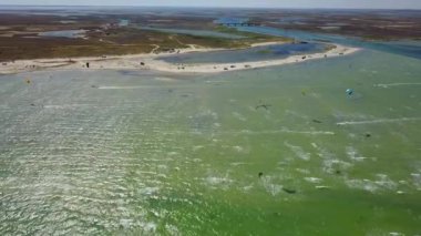 Aerial of many kiteboarders with colorful kites flying over the blue sea lagoon ride on kiteboards. Kitesurfers surf on waves with parachutes on a windy day. Drone video.