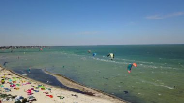 Aerial of many kiteboarders with colorful kites flying over the blue sea lagoon ride on kiteboards. Kitesurfers surf on waves with parachutes on a windy day. Drone video.