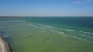 Aerial of many kiteboarders with colorful kites flying over the blue sea lagoon ride on kiteboards. Kitesurfers surf on waves with parachutes on a windy day. Drone video.
