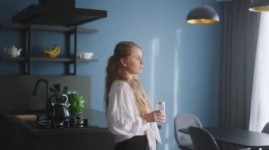 Thirsty smiling young woman standing alone in domestic kitchen drinking water from glass. Girl start a new day with healthy life habit, sipping clean mineral natural still water close up view
