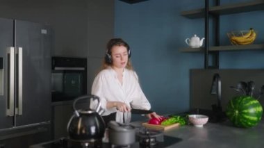 Woman in headphones making a salad of vegetables and has fun dancing at modern kitchen island. Young female cuts the greens and cooks salad. Girl cuts cucumbers and tomatoes on a chopping board.