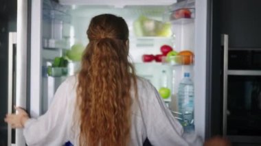 Young woman taking vegetables from fridge and has fun singing and dancing in modern minimalistic kitchen with island. Girl getting tomato from side by side refrigerator and singing to a cucumber.