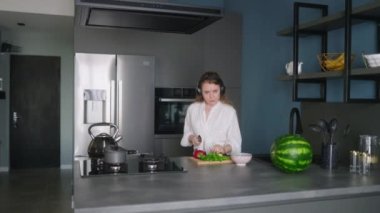 Woman in headphones making a salad of vegetables and has fun dancing at modern kitchen island. Young female cuts the greens and cooks salad. Girl cuts cucumbers and tomatoes on a chopping board.