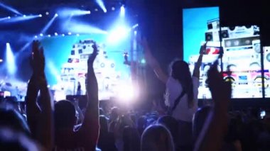 Woman sits on boyfriend shoulders clapping hands at music concert on open air fest. Crowd applause, jump, wave hands and dance at a favourite band performance on stage. Bright illumination.