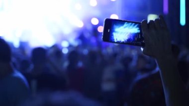People waving hands silhouettes, taking photos or recording videos of live music concert with smartphones at open air festival. Bright colorful stage lighting. Fans at a favourite band performance.