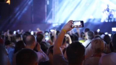 Close up of man hands silhouette taking photo or recording video of live music concert with smartphone at open air festival. Bright colorful stage lighting. Fans at the favourite band performance.