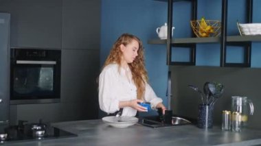 Young caucasian woman washes spoon, fork and knife under running water in sink in modern kitchen. Housewife rinsing cutlery on kitchen island. Female performing the washing-up in high-tech kitchen