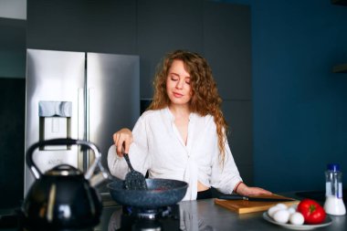 Young smiling curly caucasian woman cooks breakfast on frying pan. Young adult female making omelette on gas stove. Girl preparing her meal on modern minimalistic kitchen island. Frying