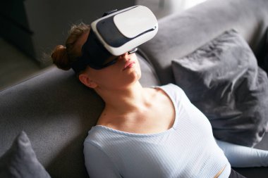 Woman in virtual reality goggles enters metaverse immersive experience via headset interface resting on sofa in minimalistic interior. Girl gaming in a cyber space. Futuristic concept.