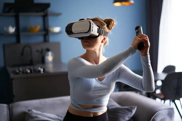 Young futuristic blonde girl wearing virtual reality headset, holding controller playing shooter video game at home. Woman playing avatar VR shooting fight in metaverse in the middle of living room.