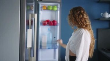 Caucasian woman opens a refrigerator and takes bottle of water, drinks cooled water. Young adult female taking sip of cold water. Girl keeps herself hydrated standing in modern kitchen closeup