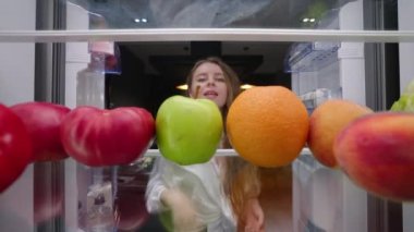 View from inside fridge - woman opens side by side fridge doors, takes fruits and vegetables, closes it. Pretty girl on fruit healthy diet. Female takes tomato, apple, peach, orange from refrigerator.