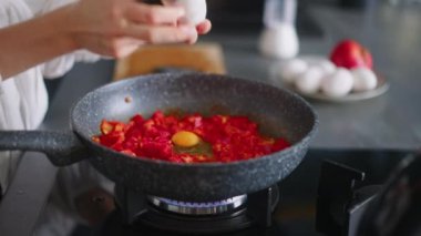 Young curly woman puts tomatos on hot pan to fry them with scrambled eggs. Young female cooking omelette on gas stove. Girl preparing her breakfast at modern minimalistic kitchen island.