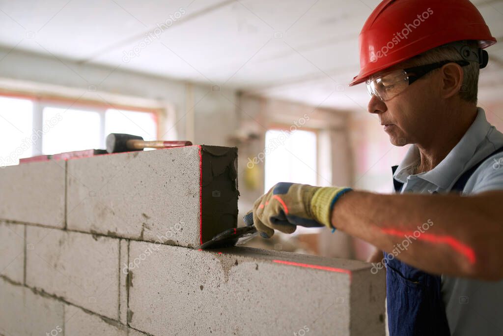 Bricklayer applies adhesive glue on autoclaved aerated concrete blocks with notched trowel. Brickwork worker contractor removes excess mortar from foam concrete doing masonry with a laser level.