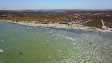 Aerial of many kiteboarders with colorful kites flying over the blue sea lagoon ride on kiteboards. Kitesurfers surf on waves with parachutes on a windy day. Drone video.